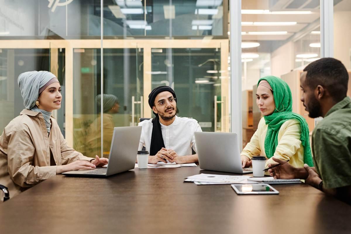 Quatre collaborateurs en hijab et casquette discutent autour de leurs ordinateurs Quatre collaborateurs en hijab et casquette discutent autour de leurs ordinateurs